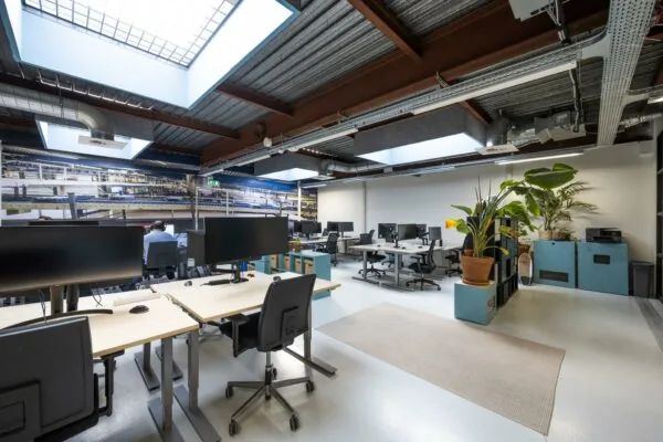 Modern, open kantoorinterieur aan de Joan Muyskenweg met bureaus, grote beeldschermen, planten en veel natuurlijk licht via dakramen.